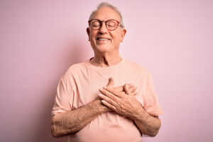 man wearing glasses standing over pink background smiling with hands on chest with closed eyes